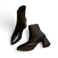 A pair of black leather ankle boots with a pointed toe and a unique flared block heel, displayed against a clean white background. One boot stands upright while the other rests on its side, highlighting its architectural silhouette and side zipper.