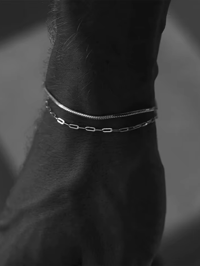 Bracciale a maglia graffetta indossato al polso insieme a una catena piatta, foto in bianco e nero